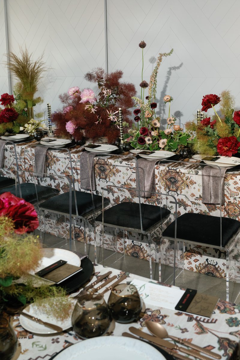 A long dining table is meticulously set with a rich, patterned tablecloth, modern place settings, and elaborate floral a