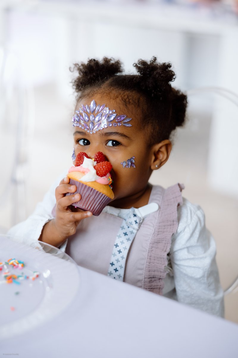 A young child with face gems, including a purple tiara design on her forehead, holds a cupcake topped with raspberries a