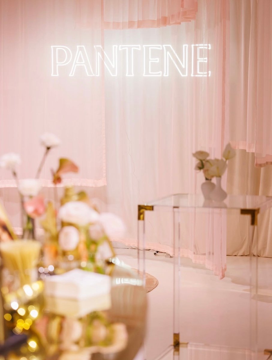This photo captures a stylish event space featuring soft pink drapes and a prominent 'PANTENE' neon sign. In the foregro