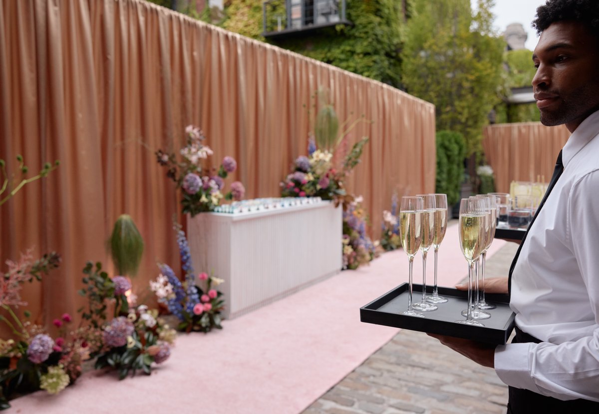 A server in a white shirt and black tie carries two trays of champagne flutes filled with sparkling wine, walking past a
