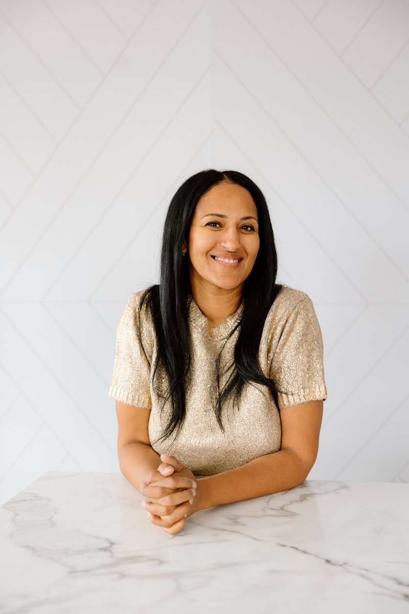 A smiling woman with long dark hair, wearing a shimmering gold top, sits at a white marble table with her hands clasped.