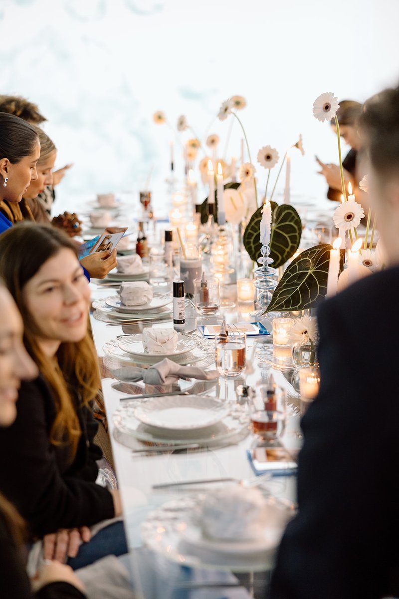 Guests are seated at a long, elegantly set dinner table, adorned with candles, white flowers, and large green leaves. Th