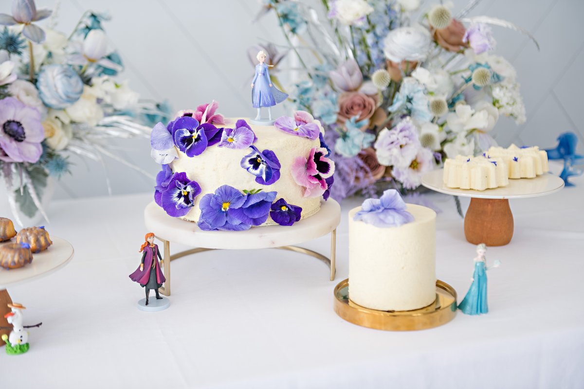 A close-up shot of a round cake, likely vanilla with buttercream frosting, adorned with vibrant purple and pink pansies.
