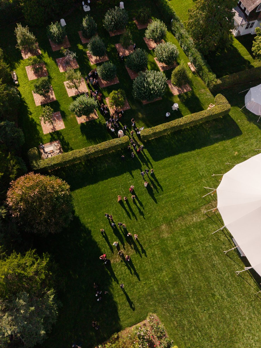 An aerial view captures guests gathering in a beautifully manicured garden. People are seen walking along paths and cong