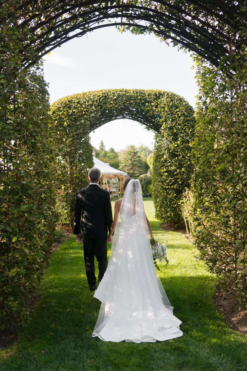 A couple, likely a bride and groom, walks hand-in-hand down a grassy path, framed by lush green archways. The path leads