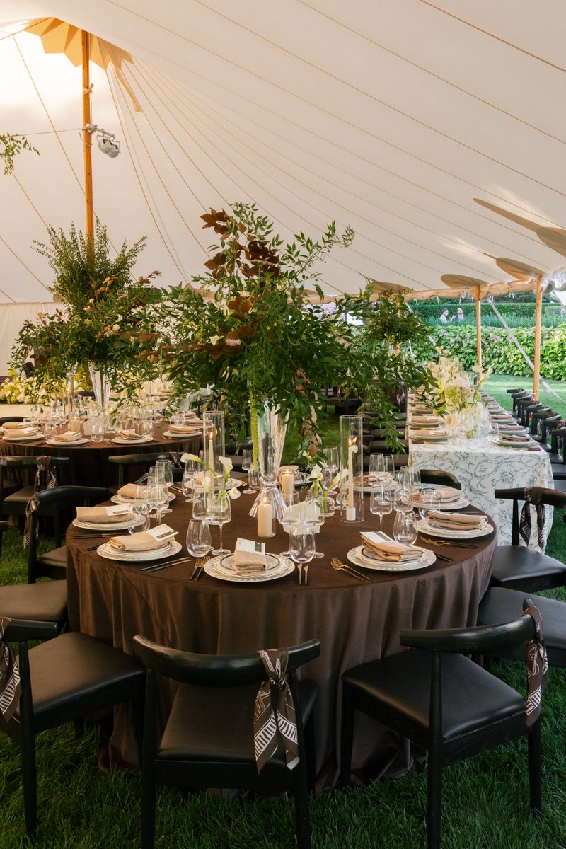 An elegantly set round dinner table is featured under a large white tent, surrounded by lush green grass. The table is a