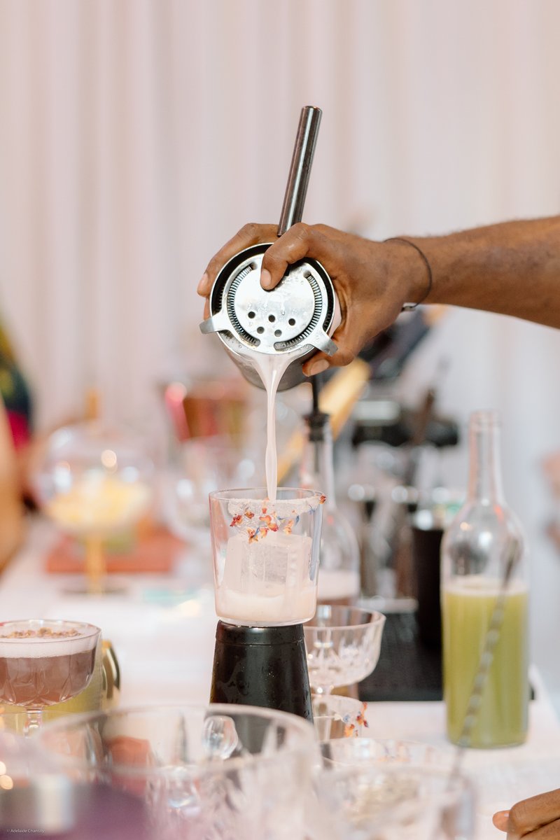 A bartender's hand is actively pouring a light pink cocktail from a shaker into a glass filled with ice cubes. The glass