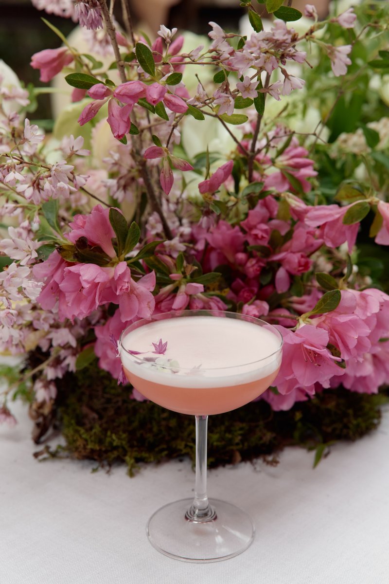 A beautifully presented pink cocktail with a foam top and delicate floral garnish sits on a white tablecloth, set agains