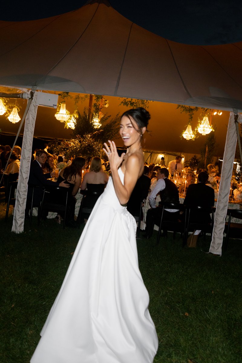 A joyful woman in a white gown, likely a bride, smiles and gestures towards the camera. In the background, guests are se