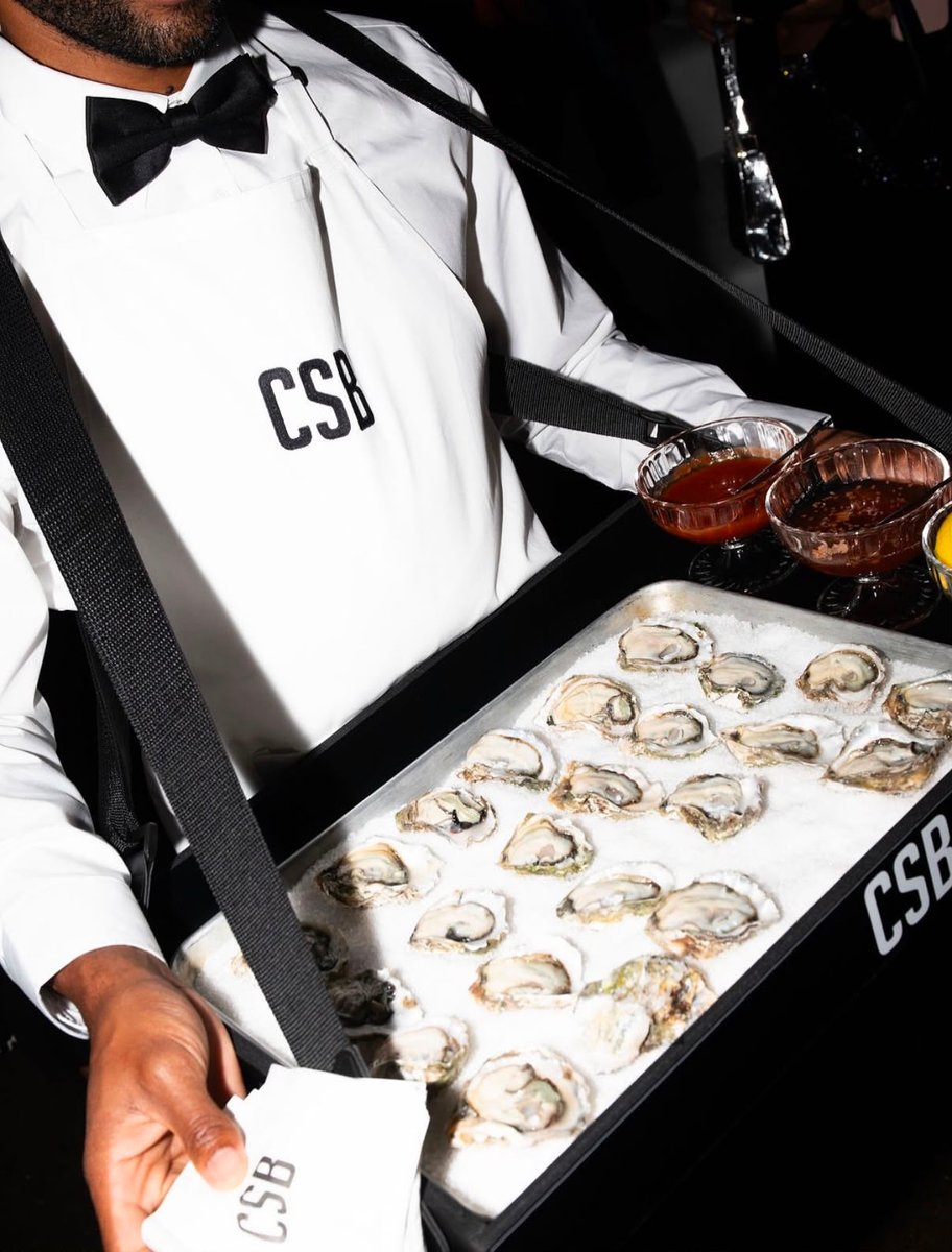 A server, dressed in a white shirt, black bow tie, and suspenders, carries a portable oyster station. The station holds 