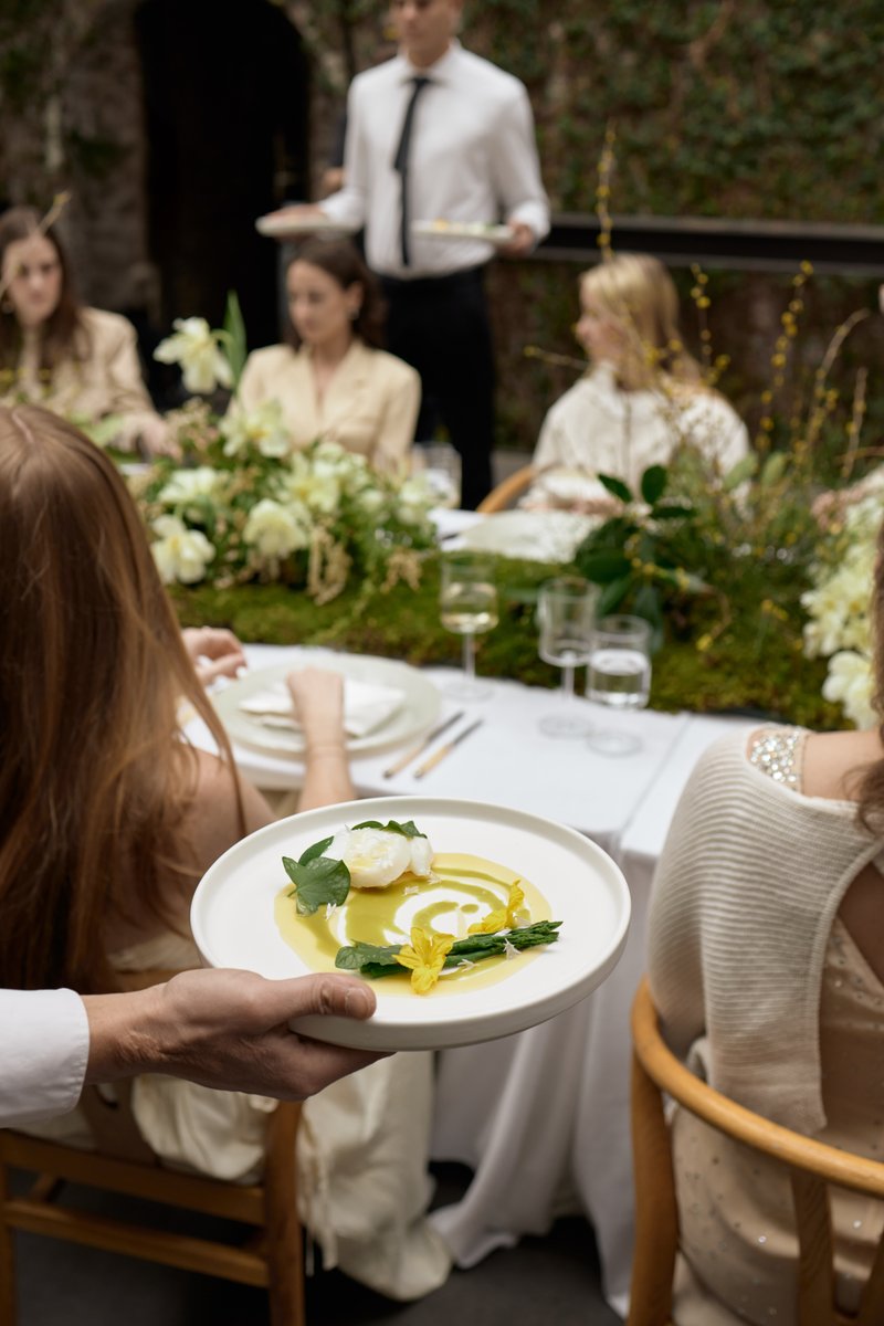 A server presents a meticulously plated dish to guests seated at a long, elegantly decorated table. The dish features a 