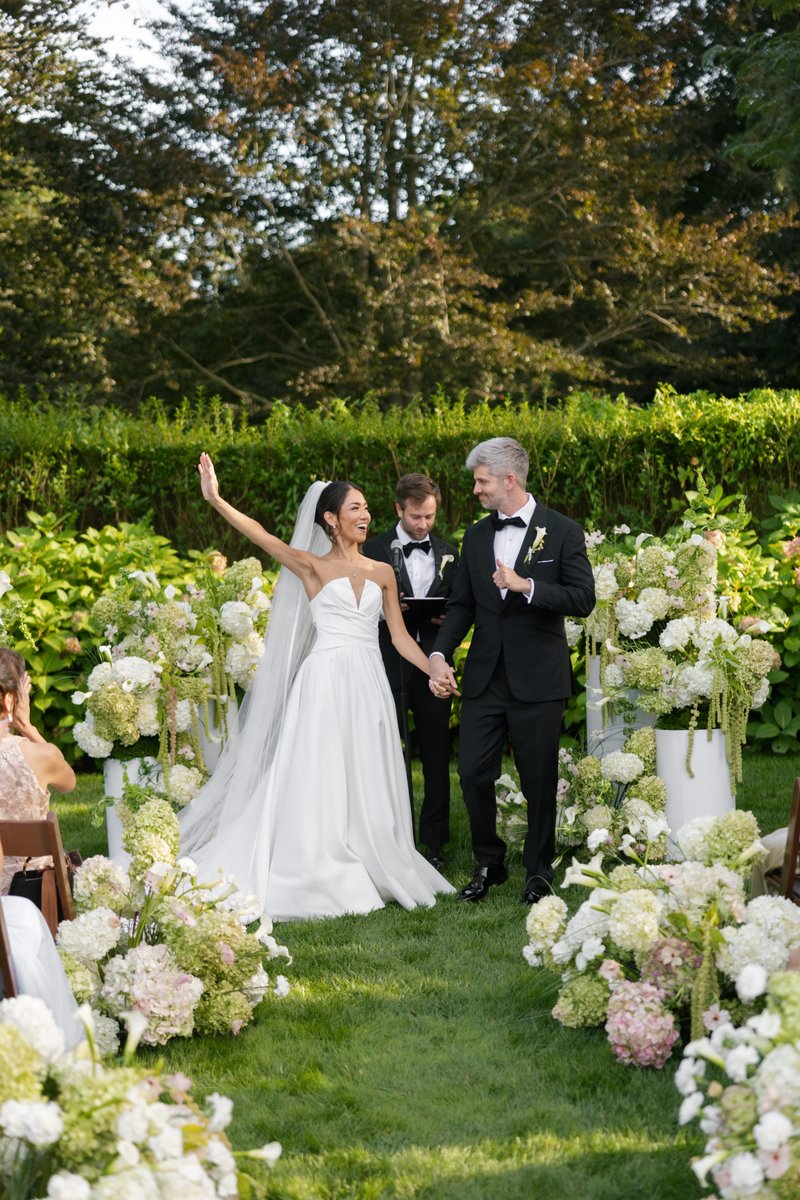 A newlywed couple, a bride in a white gown and a groom in a black tuxedo, walk hand-in-hand down a grassy aisle after th