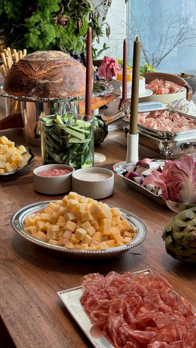 A lavish spread of charcuterie, cheeses, and fresh vegetables is presented on a wooden table. Various silver platters an