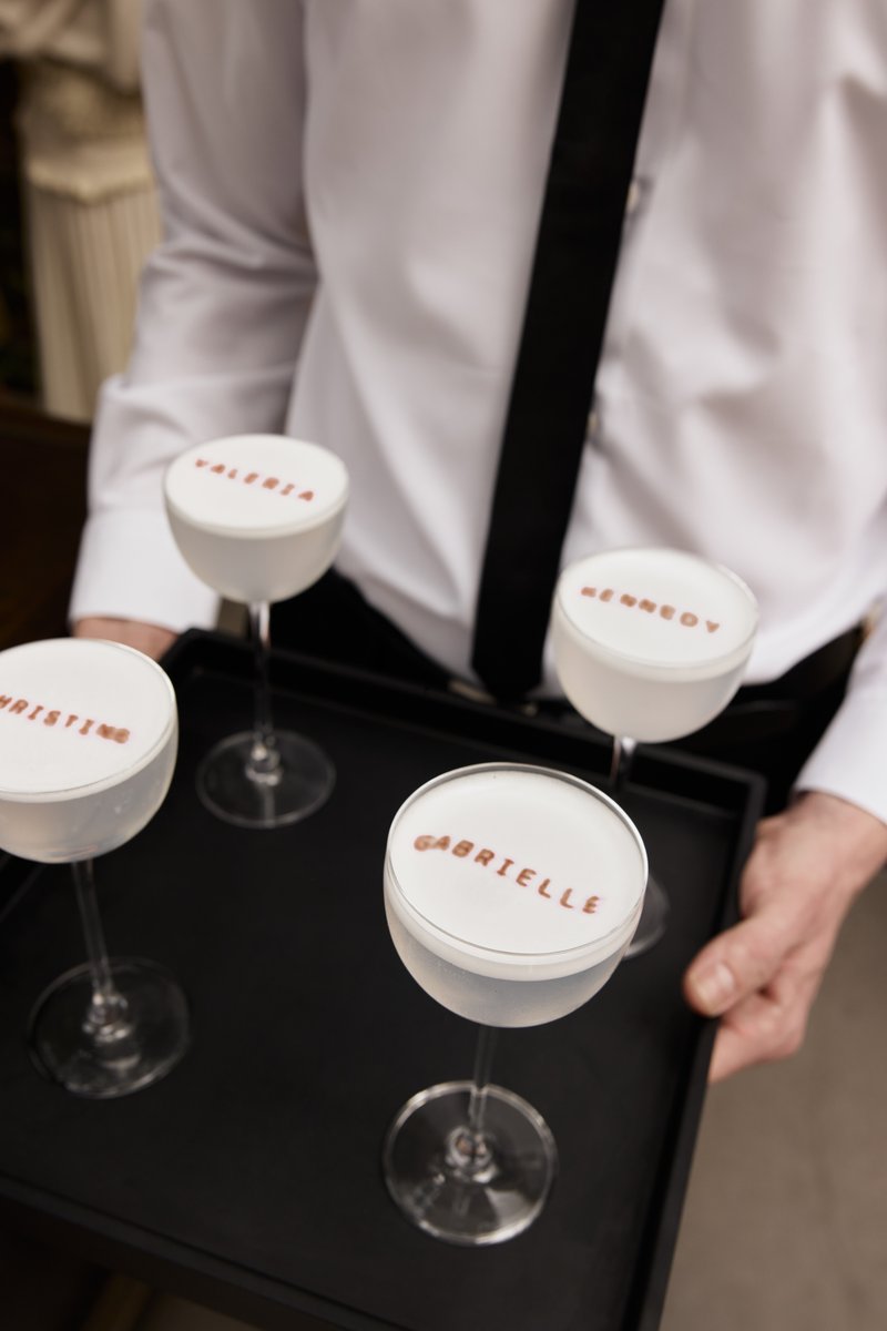A server, dressed in a white shirt and black tie, holds a black tray with four personalized cocktails. Each cocktail fea