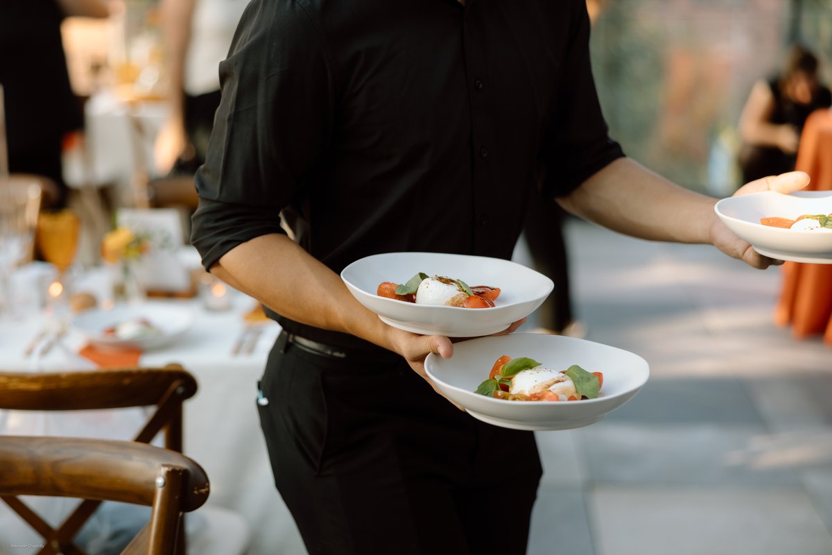 A server in a black uniform is expertly carrying multiple plates of an appetizer, likely a Caprese-style salad, through 