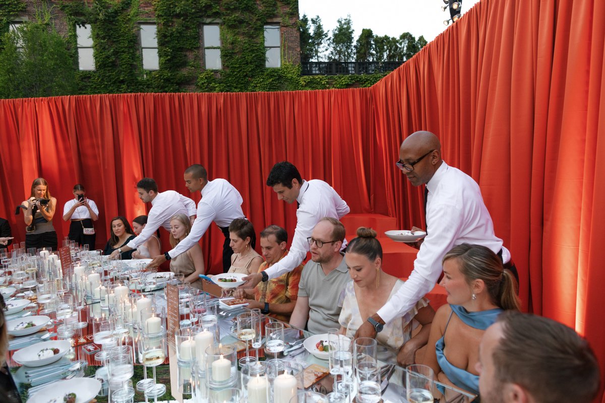 Servers in white shirts are attentively serving plated dishes to guests seated at a long, candlelit table outdoors. The 