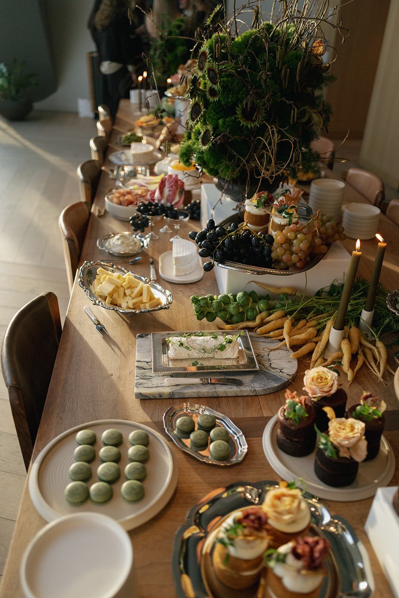 A beautifully arranged grazing table is laden with an assortment of cheeses, fresh fruits, vegetables, and desserts. Two