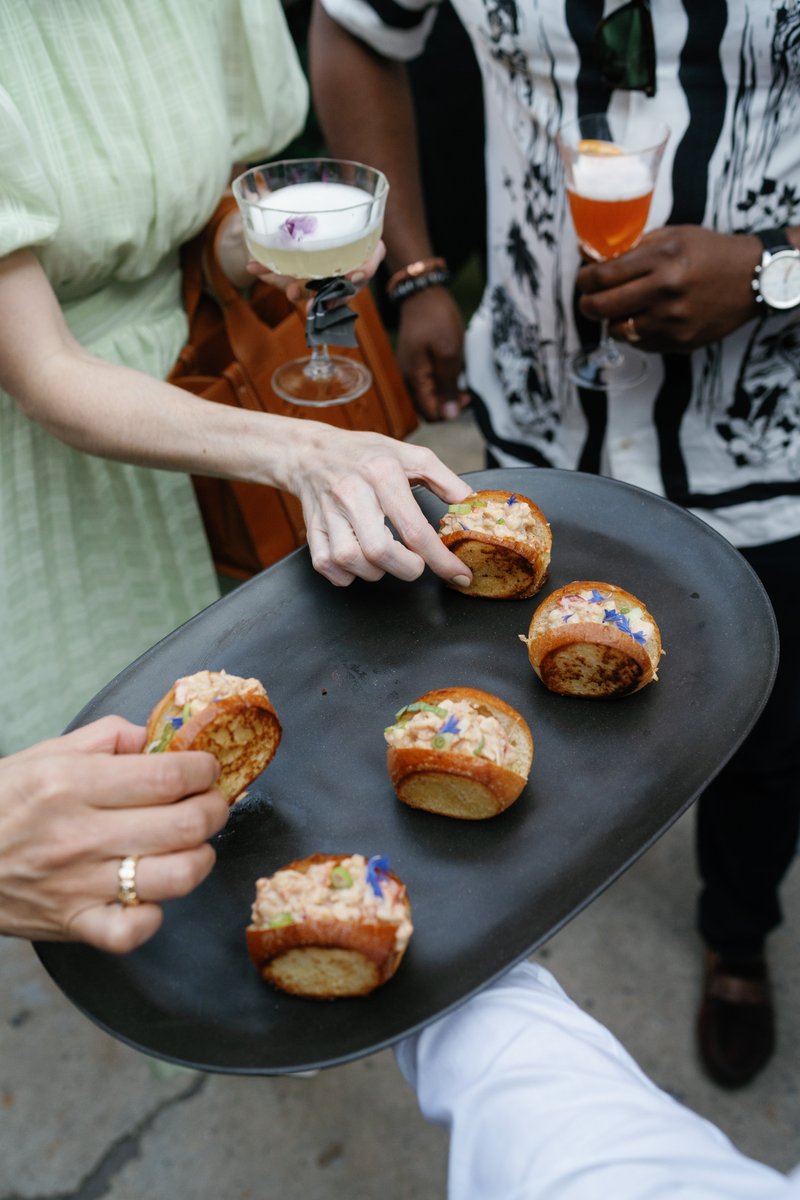 Guests are seen taking appetizers from a black serving tray held by a server, while also holding cocktails. The mood is 