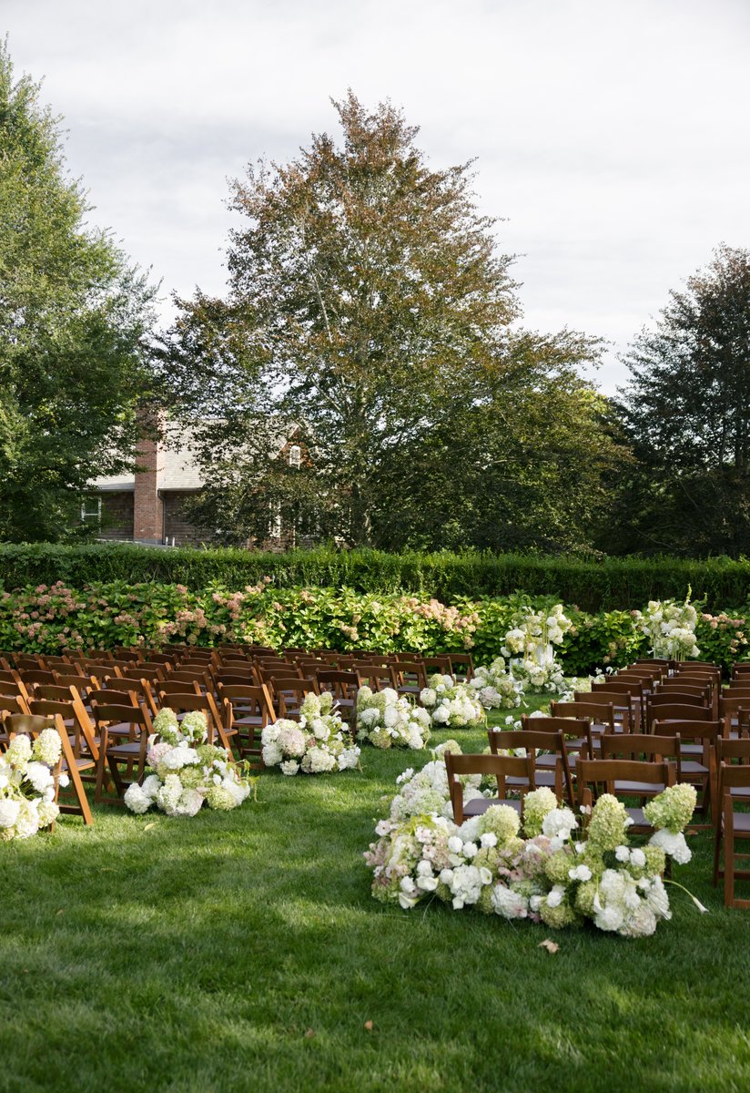 An elegant outdoor ceremony space is prepared on a lush green lawn. Rows of dark wooden chairs face a hedge, with beauti