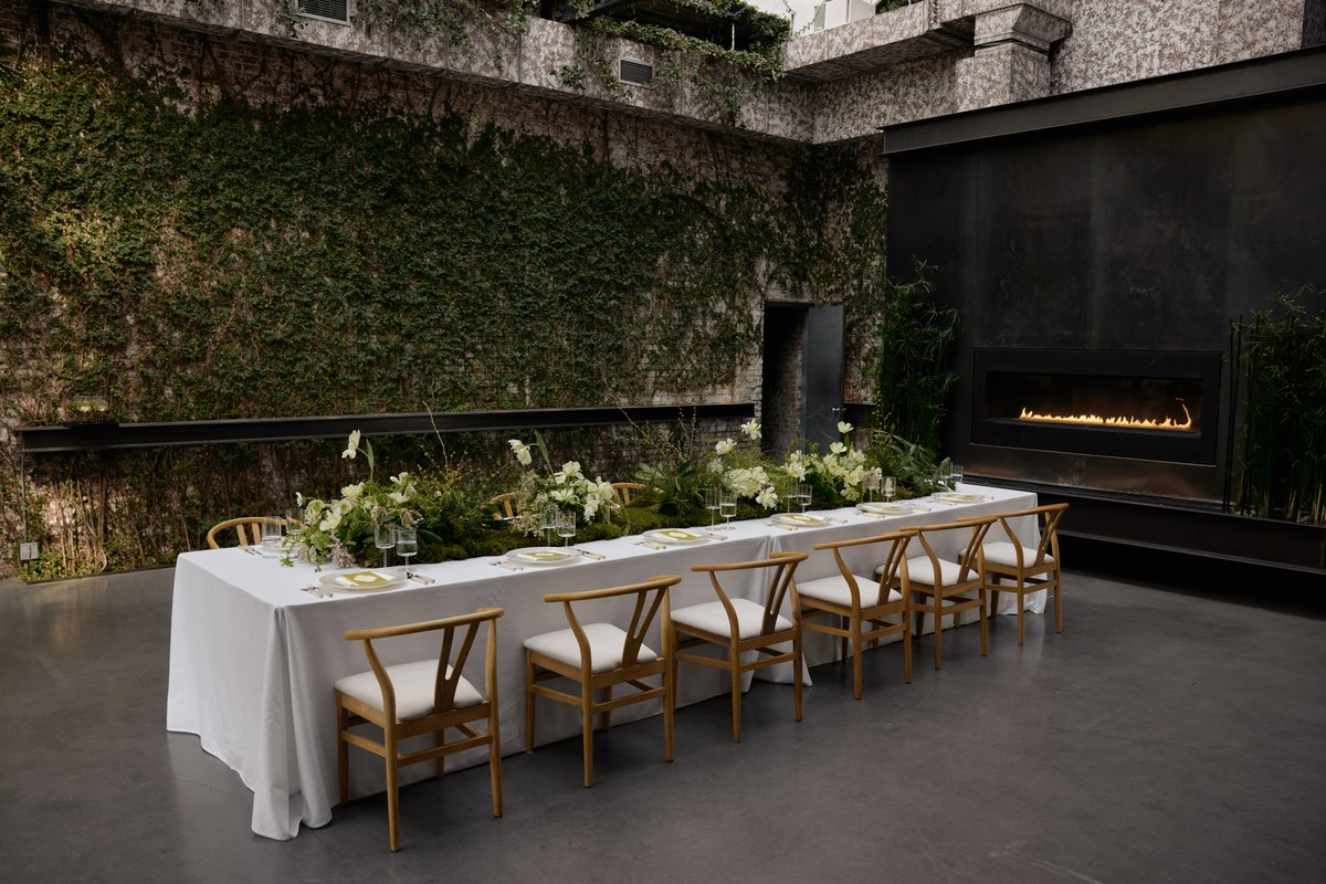 A long dining table, elegantly set with white linens and modern wooden chairs, is adorned with lush green moss and white