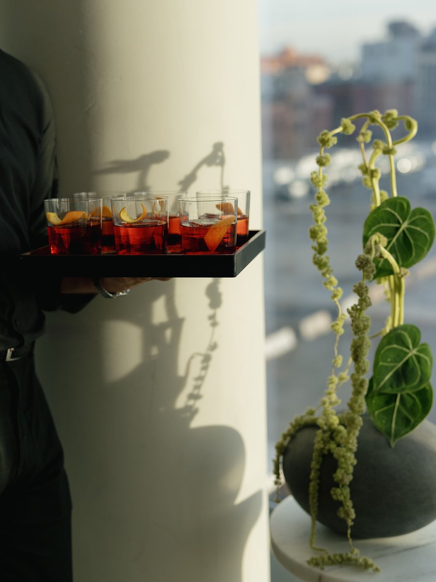 A server carries a tray of vibrant red cocktails, each garnished with an orange peel, through an event space. In the for