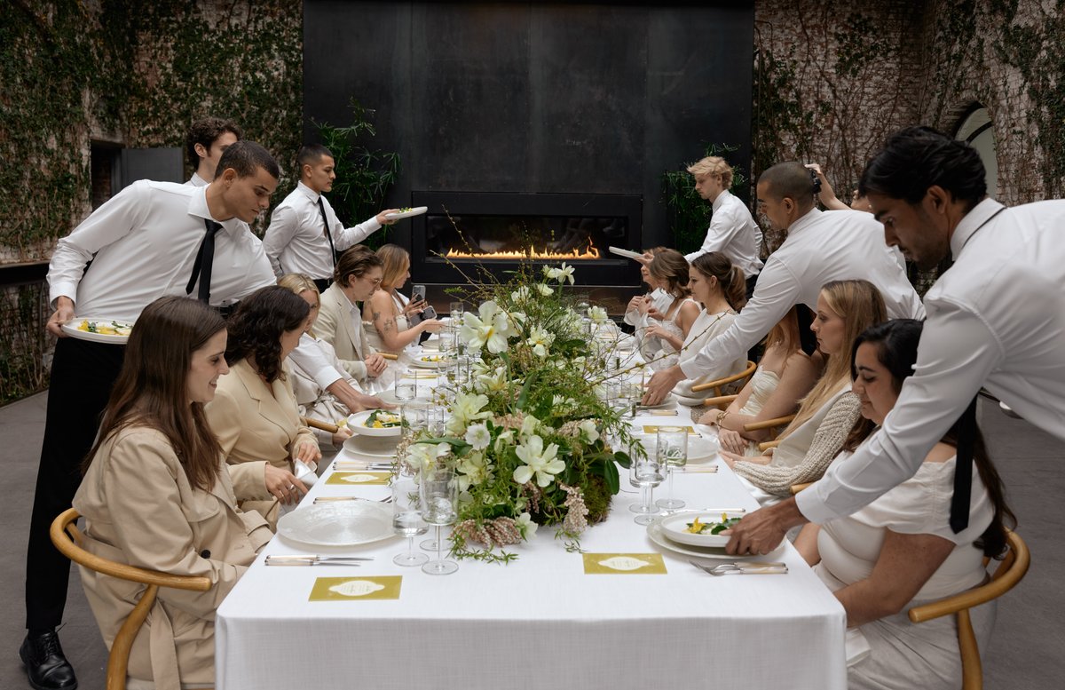 Servers in white shirts and black ties are attentively serving plated dishes to guests seated at a long, elegantly set t