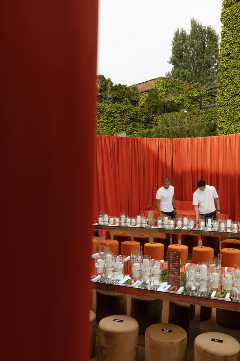 Two staff members are meticulously setting up long tables adorned with numerous unlit candles and place settings, agains