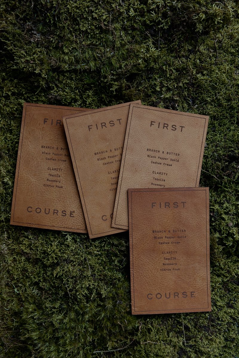Several rustic, leather-like menu cards are fanned out and displayed on a bed of vibrant green moss. The cards detail a 