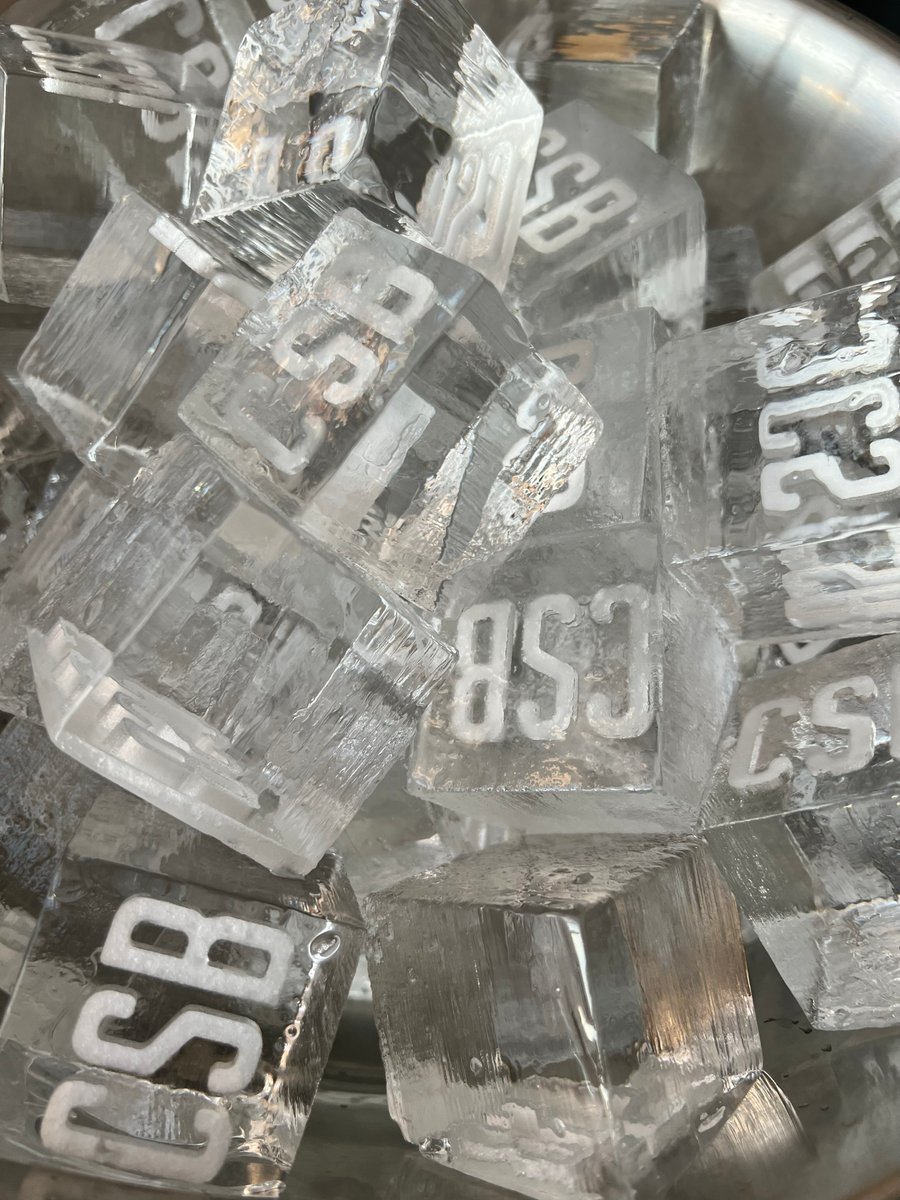 A close-up shot of custom ice cubes, each featuring the letters "CSB" embedded within, resting in a metal bucket. The ic