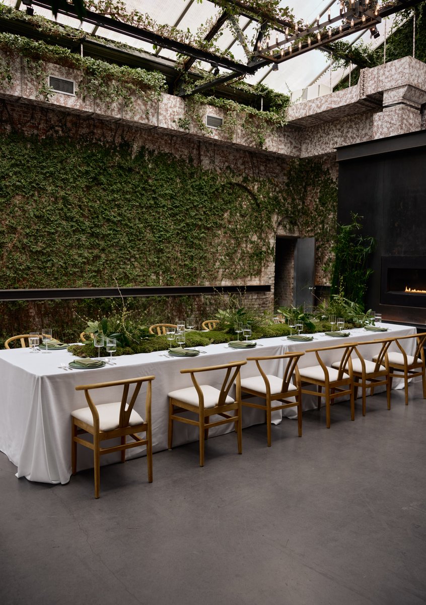 A long dining table, elegantly set with white linens and natural green moss and foliage as a centerpiece, is positioned 