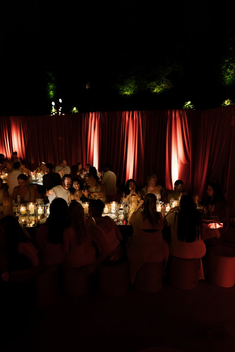 Guests are seated at a long, candlelit table, enjoying an intimate dinner or event. The setting features dramatic red dr
