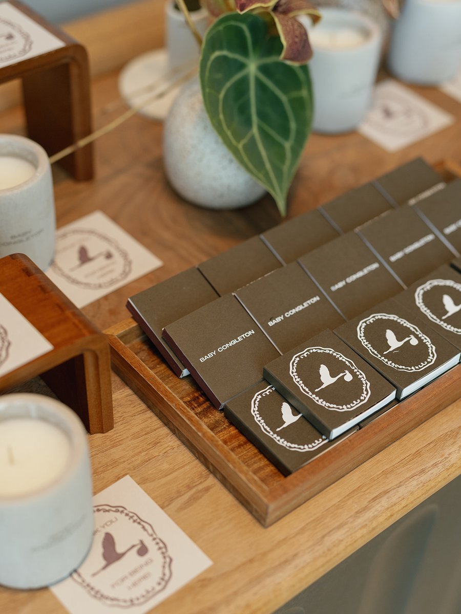 A close-up shot of event favors, likely matchbooks, arranged on a wooden tray. They feature a stork logo and the text 'B