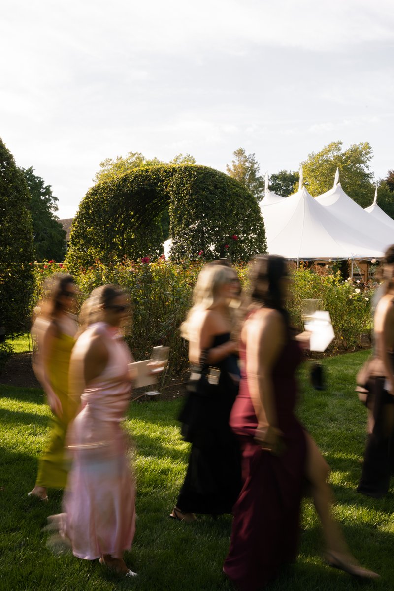 Several guests in formal attire are captured in motion, walking across a sunlit grassy area at an outdoor event. In the 