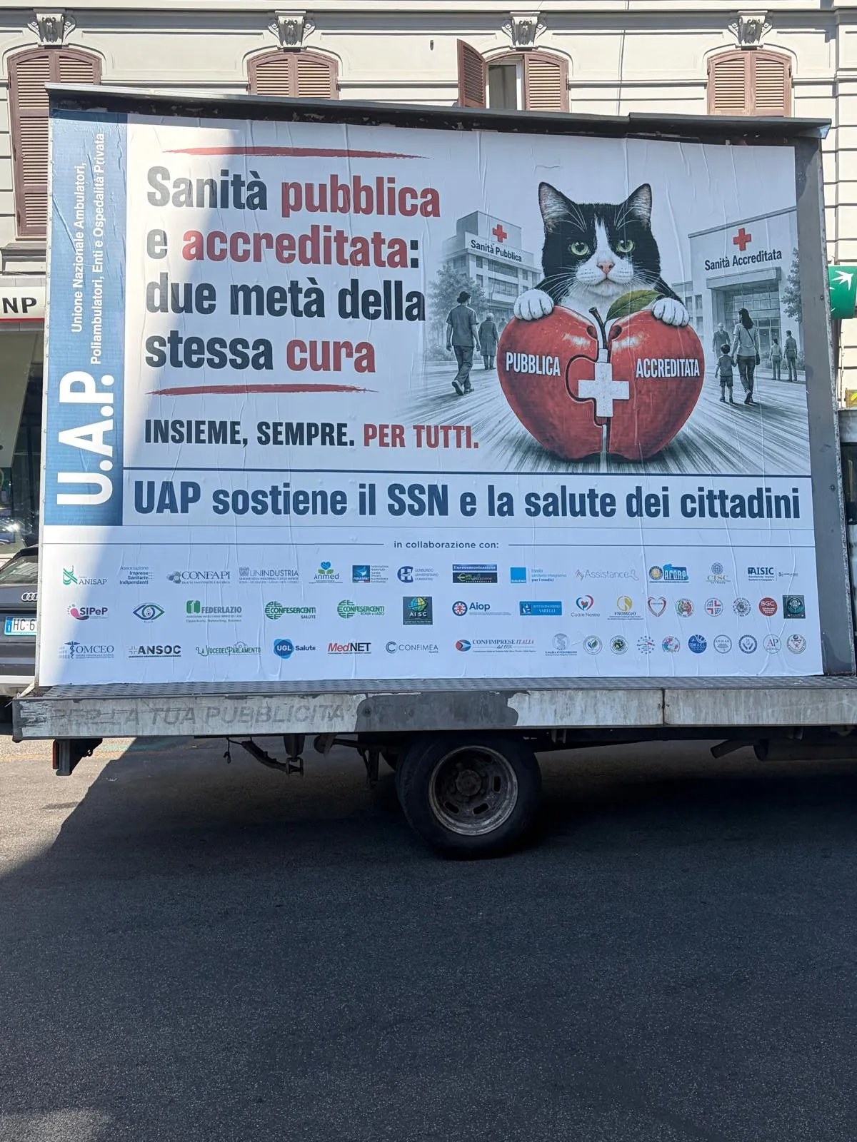 L'immagine mostra un grande cartellone pubblicitario su un camion, che promuove la sanità pubblica e accreditata in Italia. Il cartellone è decorato con un'immagine di un gatto che tiene in una zampa una sferetta rossa con il simbolo della croce rossa, sottolineando la tematica della salute.