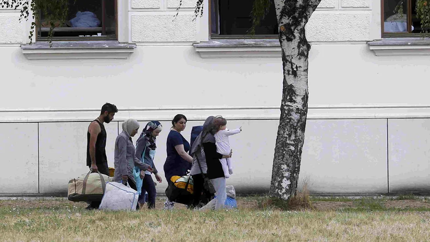 L'immagine mostra un gruppo di persone che camminano lungo un marciapiede, trasportando bagagli. Sono visibili diverse donne e uomini, alcuni dei quali indossano veli. Sullo sfondo si intravedono finestre di un edificio.