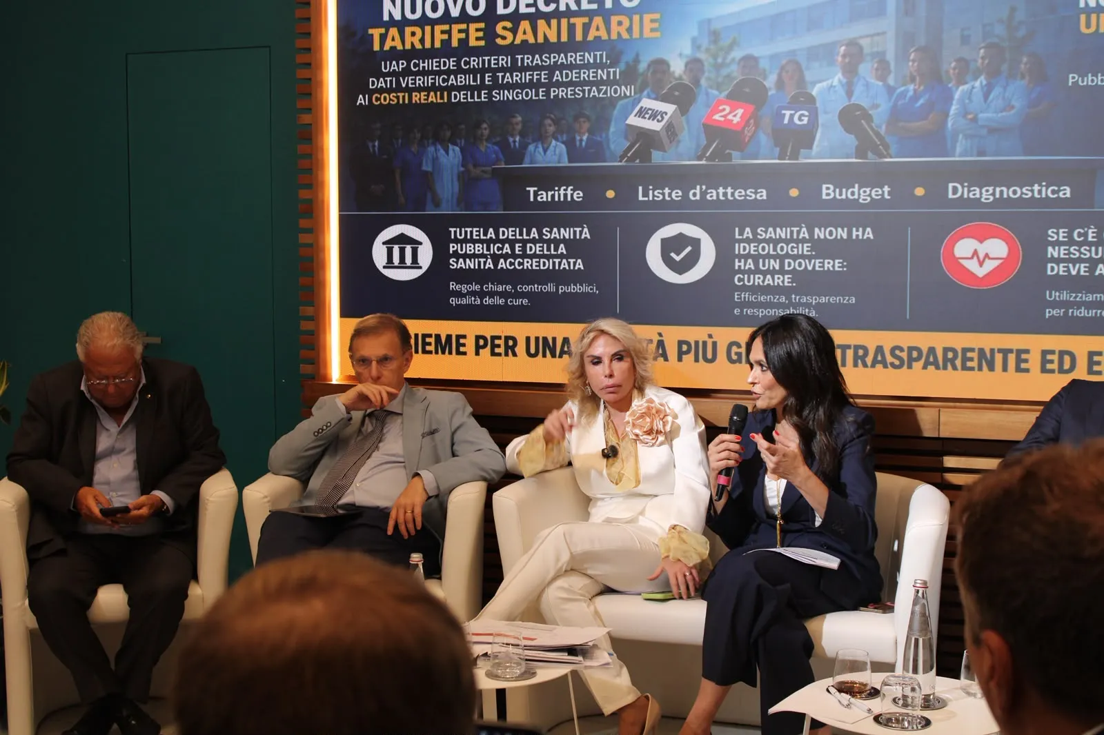 L'immagine mostra un panel di discussione pubblica su temi sanitari. Al centro, una relatrice in abbigliamento elegante parla al microfono, mentre altri partecipanti l'ascoltano con attenzione. Sullo sfondo, è visibile un banner informativo relativo a tariffe sanitarie e trasparenza.