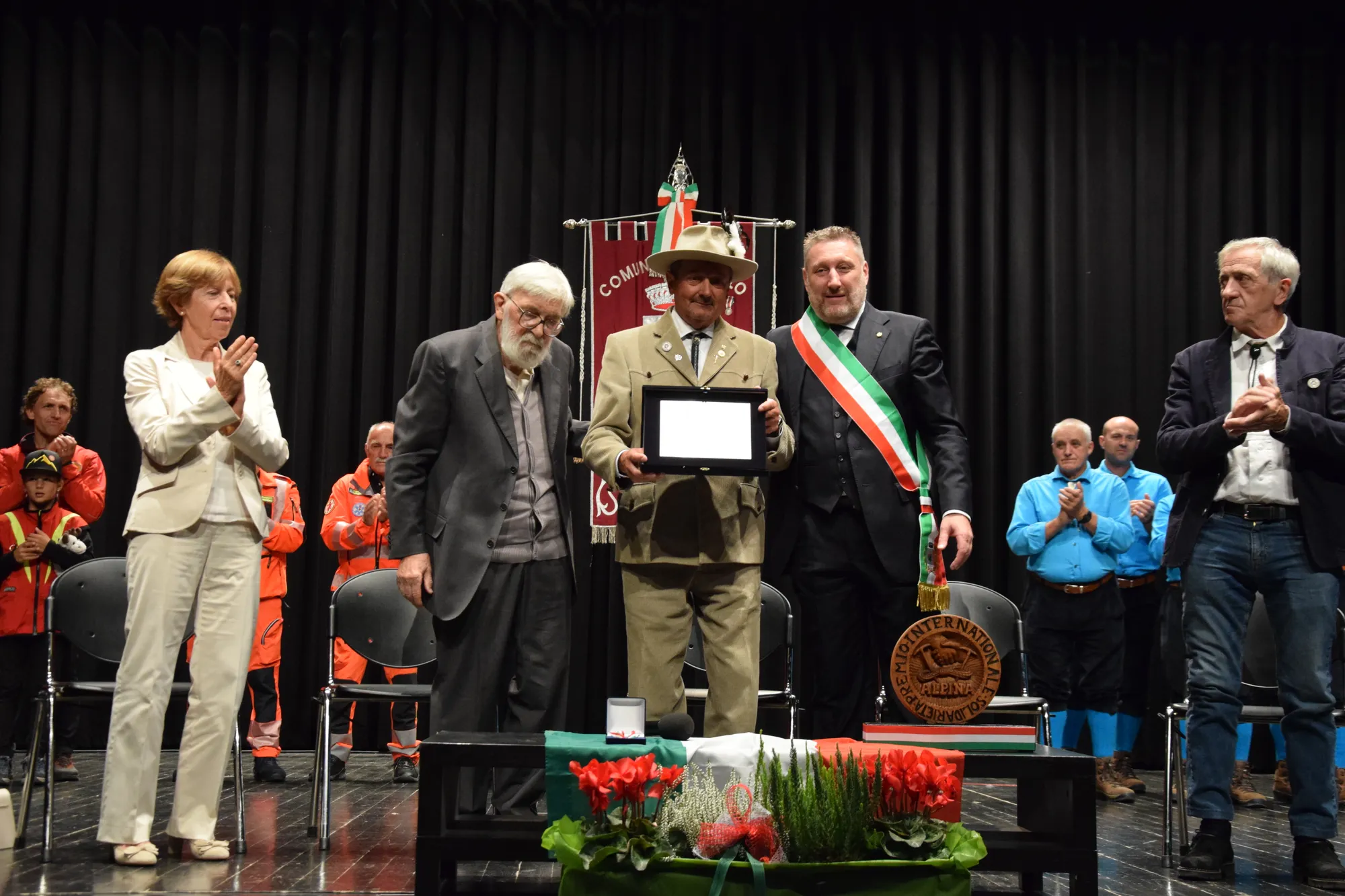 L'immagine mostra una cerimonia di premiazione con diverse persone sul palco. Al centro, un uomo in uniforme tiene un premio, circondato da altri partecipanti che applaudono. Sullo sfondo, si notano simboli e colori legati a un evento ufficiale.
