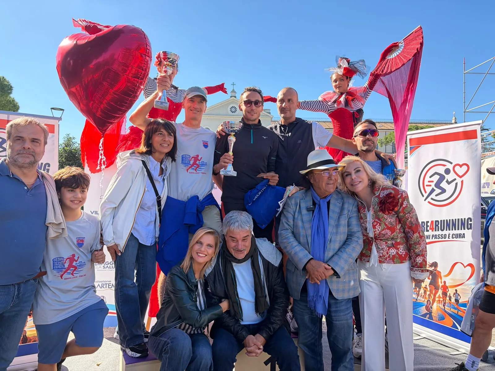 L'immagine ritrae un gruppo di persone festose in occasione di un evento, probabilmente una competizione sportiva o una manifestazione di beneficenza. Alcuni partecipanti sorreggono trofei e ci sono decorazioni a tema, come cuori e palloncini rossi, che conferiscono un'atmosfera vivace e gioiosa.