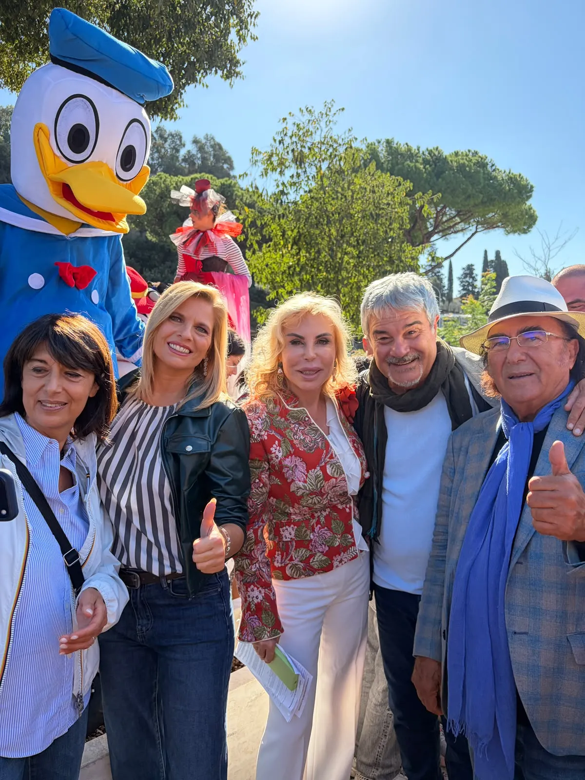 L'immagine mostra un gruppo di persone che sorridono e posano insieme per una foto. Sullo sfondo, si vedono sia figure vestite in costumi festivi che un grande personaggio animato, presumibilmente rappresentante un famoso papero. L'atmosfera appare festosa e gioiosa.
