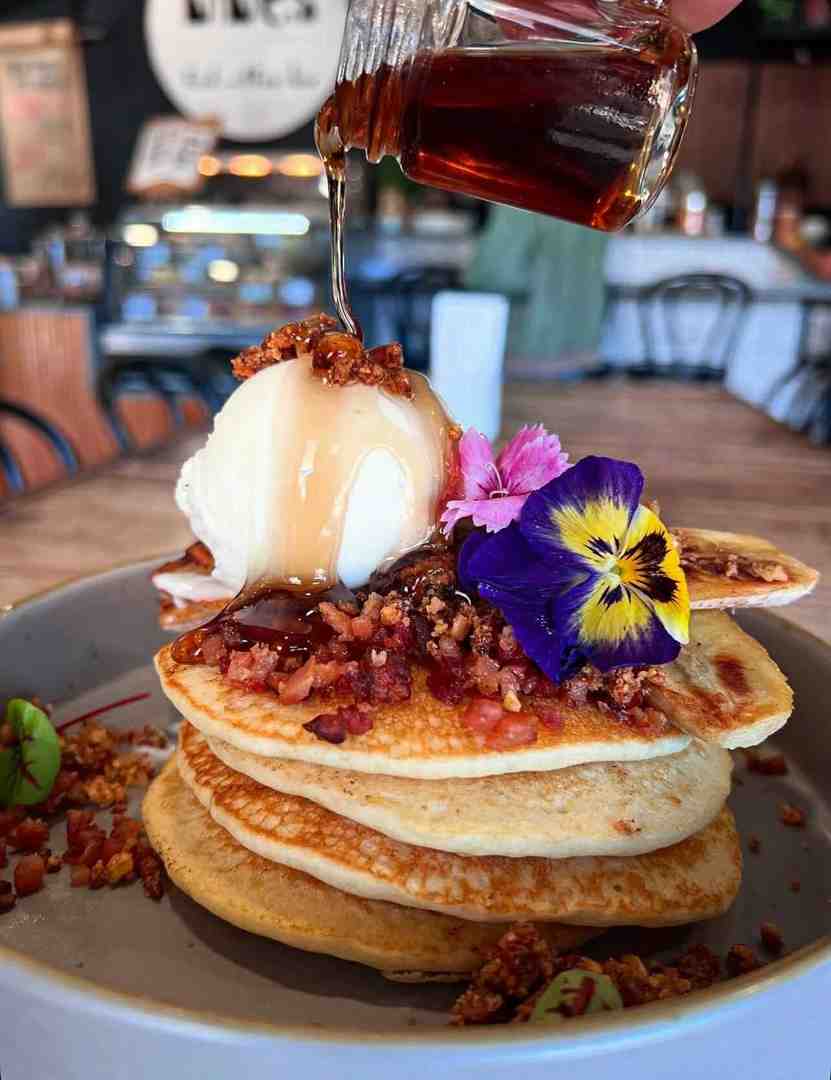 Indulgent stack of fluffy pancakes with ice cream, crispy bacon pieces, and edible flowers, being drizzled with maple syrup; a popular all day brunch choice at The Nines Indulgent stack of fluffy pancakes with ice cream, crispy bacon pieces, and edible flowers, being drizzled with maple syrup; a popular all day brunch choice at The Nines