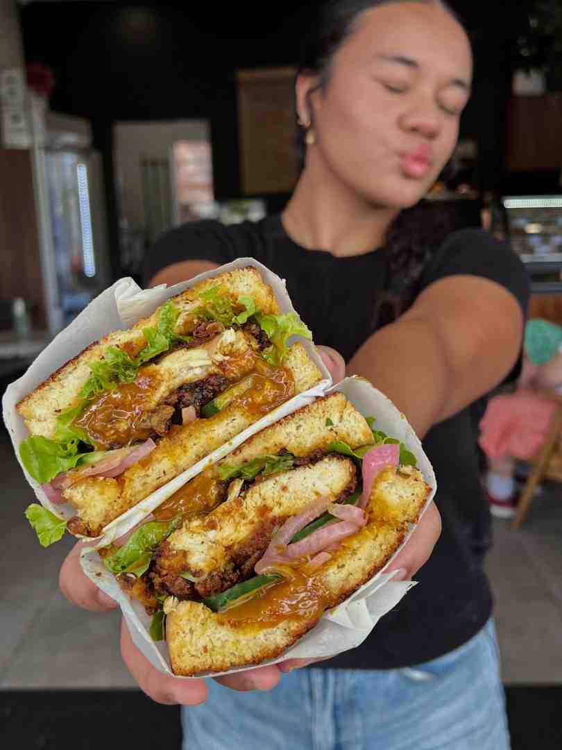 A vibrant image of a woman presenting a giant, fried chicken toasted sandwich with pickled onion and sauce. Excellent lunch specials Sunshine Coast. A vibrant image of a woman presenting a giant, fried chicken toasted sandwich with pickled onion and sauce. Excellent lunch specials Sunshine Coast.
