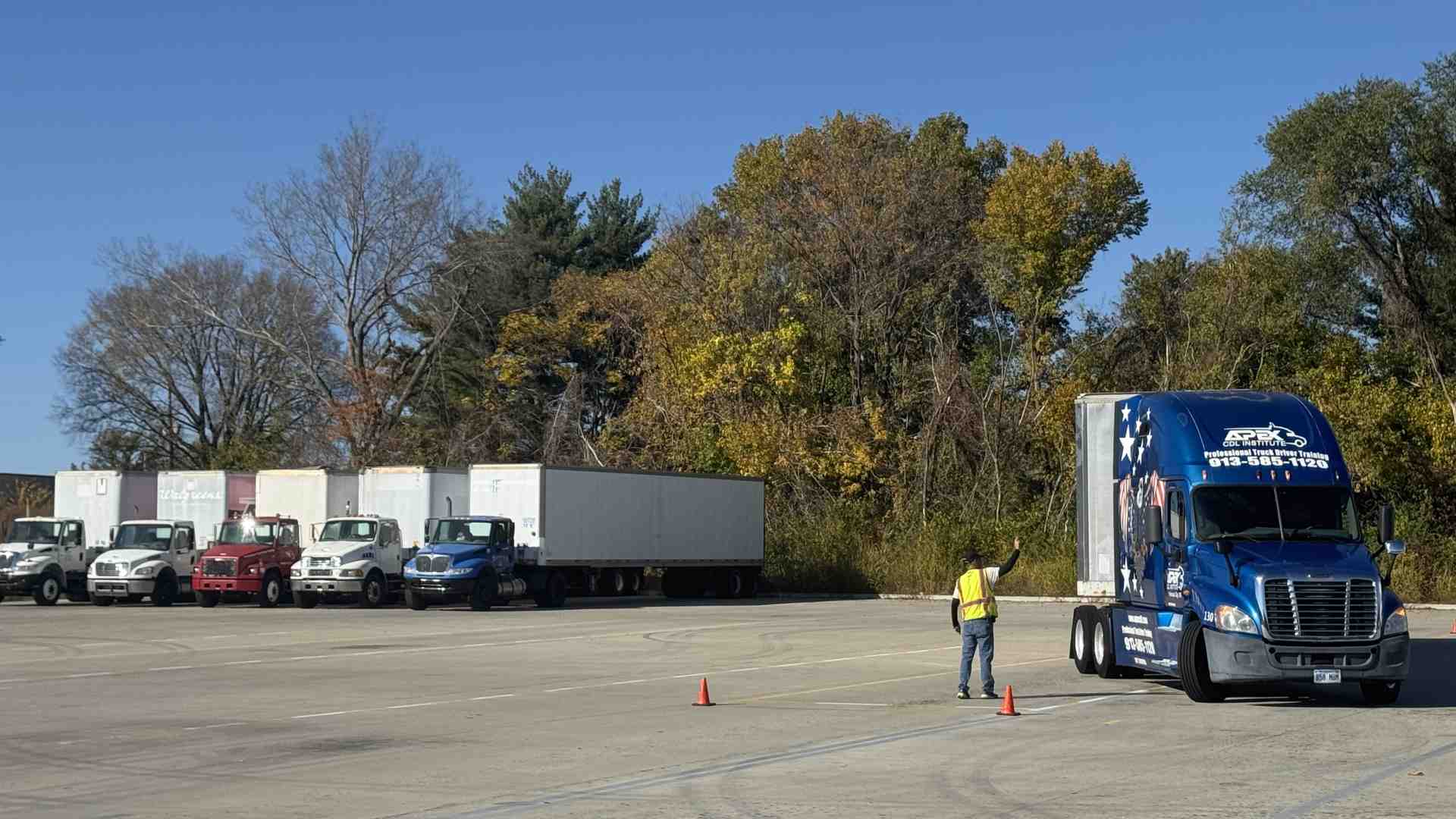 Student maneuvers an APEX truck as instructor guides in training lot with parked trucks and fall trees in background.