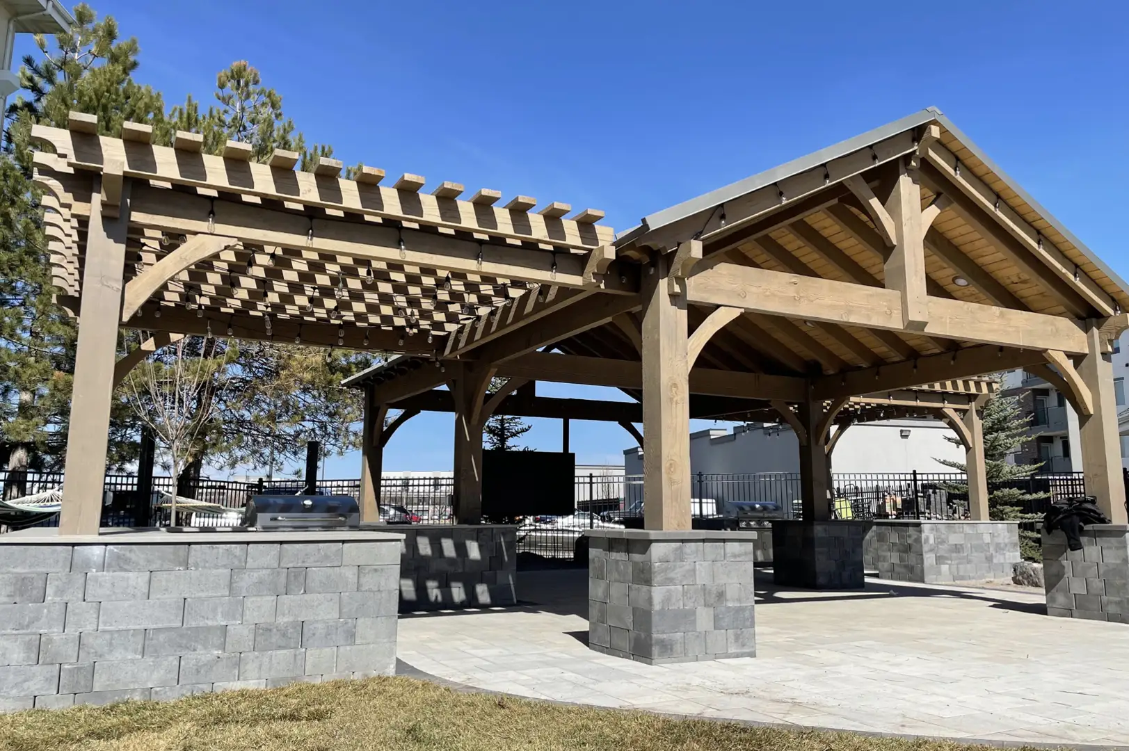 Timber Frame Outdoor Kitchen Pergola Large timber frame outdoor kitchen pergola with open rafters, stone base columns, and built in grill area under clear blue sky.