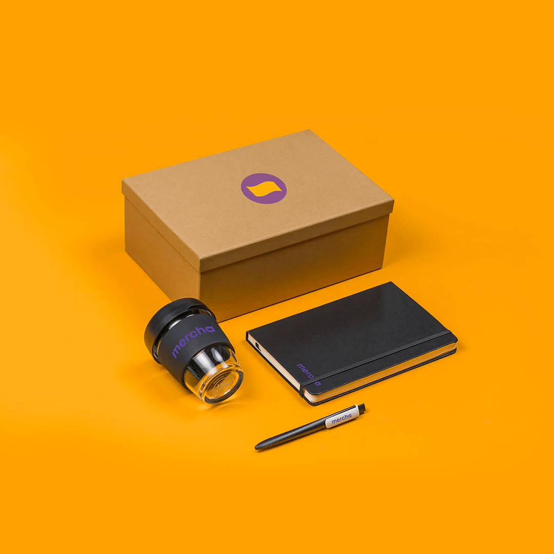 A curated set of premium promotional items from Mercha, including a kraft-coloured box, black notebook, pen, and reusable glass coffee cup with a black silicone band and lid.