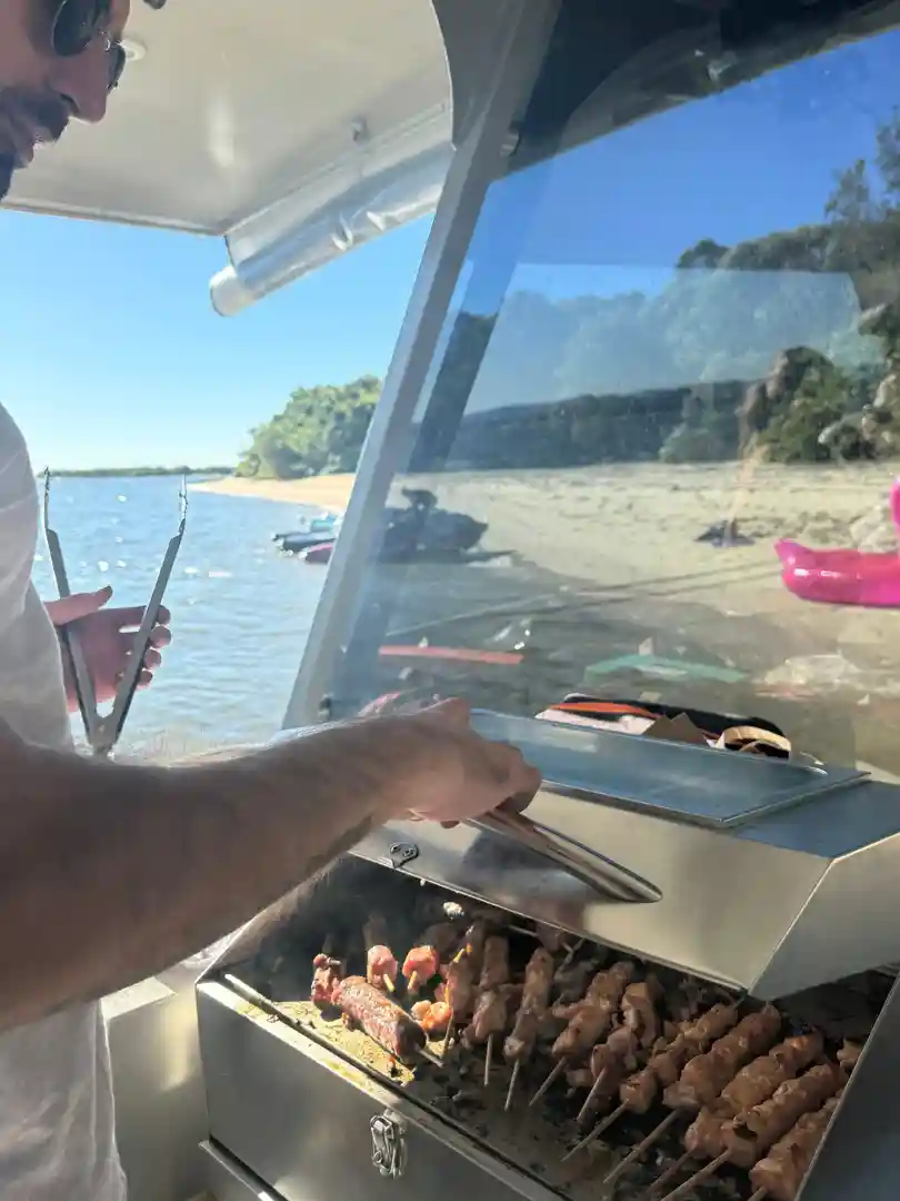 Man grilling skewers and sausages on a BBQ pontoon on the Gold Coast, with a sandy beach and watercraft in the background - hire jet ski Gold Coast