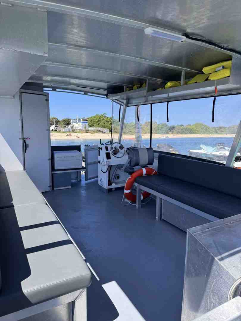Interior view of a modern pontoon boat, showing the helm, grey bench seating, a life ring, and a clear view of a sunny beach and water - Pontoon boat for beginners Interior view of a modern pontoon boat, showing the helm, grey bench seating, a life ring, and a clear view of a sunny beach and water - Pontoon boat for beginners