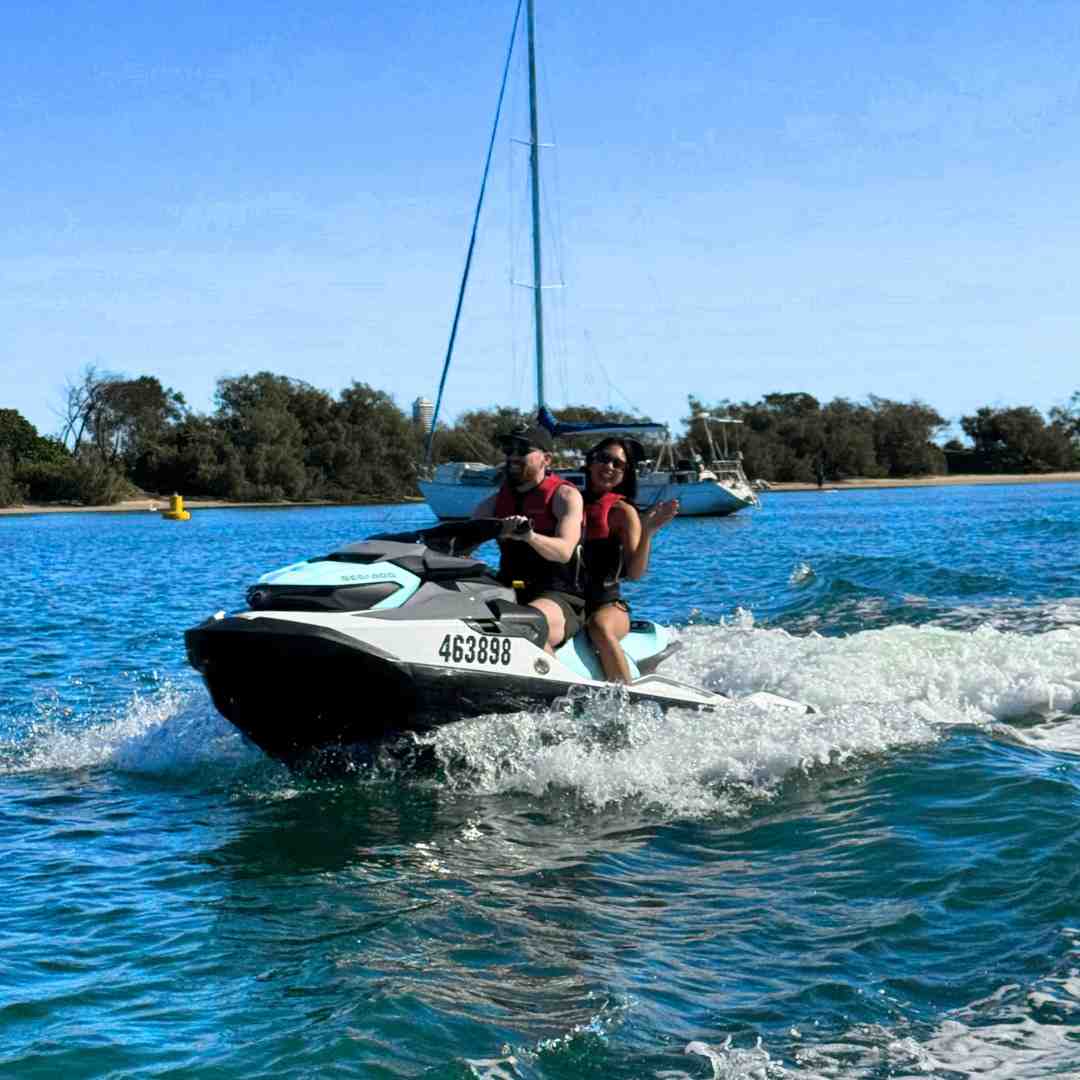Excited couple enjoying a fast ride on a jet ski near a sailboat on a sunny day - hire jet ski Gold Coast