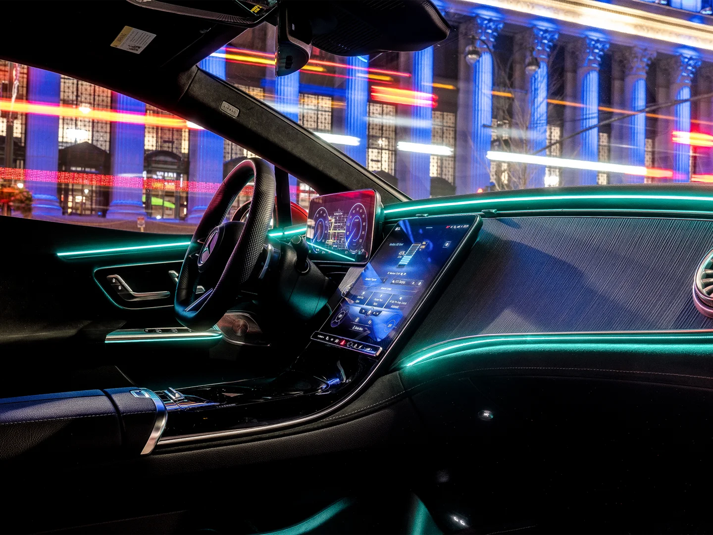 Interior cockpit view of a Mercedes-Benz featuring vibrant cyan ambient lighting and a large digital touchscreen infotainment system.