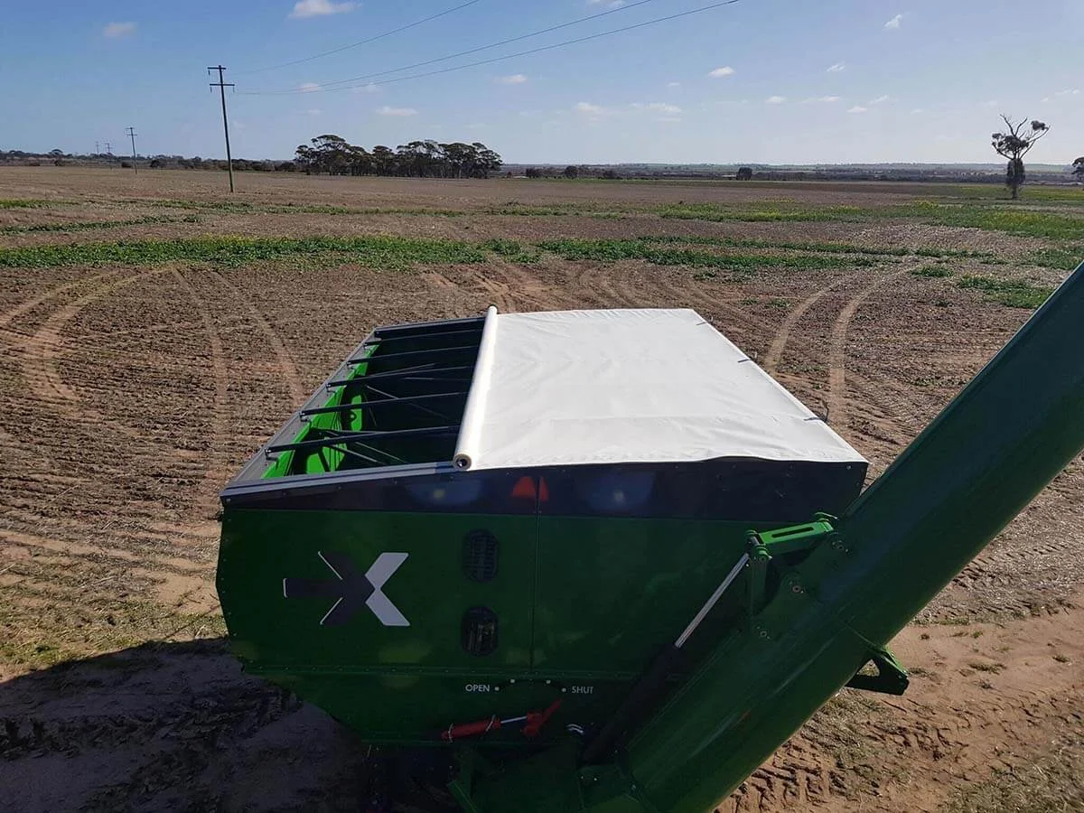 A green agricultural trailer in a field with a white side-to-side rollover tarp system, partly rolled. A green agricultural trailer in a field with a white side-to-side rollover tarp system, partly rolled.