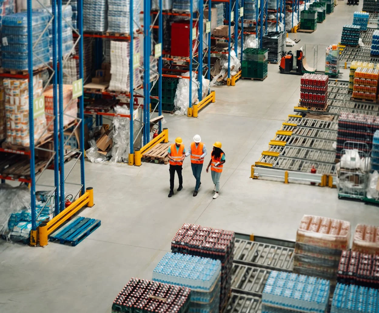 Three workers in reflective vests and hard hats walk through a massive warehouse filled with pallets